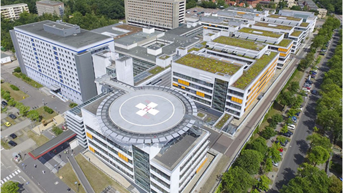 Luftaufnahme eines großen Krankenhauskomplexes mit mehreren miteinander verbundenen Gebäuden, einem Hubschrauberlandeplatz mit rotem Kreuz, begrünten Dächern und umliegenden Bäumen und Straßen.