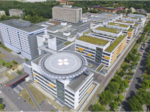 Luftaufnahme eines großen Krankenhauskomplexes mit mehreren miteinander verbundenen Gebäuden, einem Hubschrauberlandeplatz mit rotem Kreuz, begrünten Dächern und umliegenden Bäumen und Straßen.