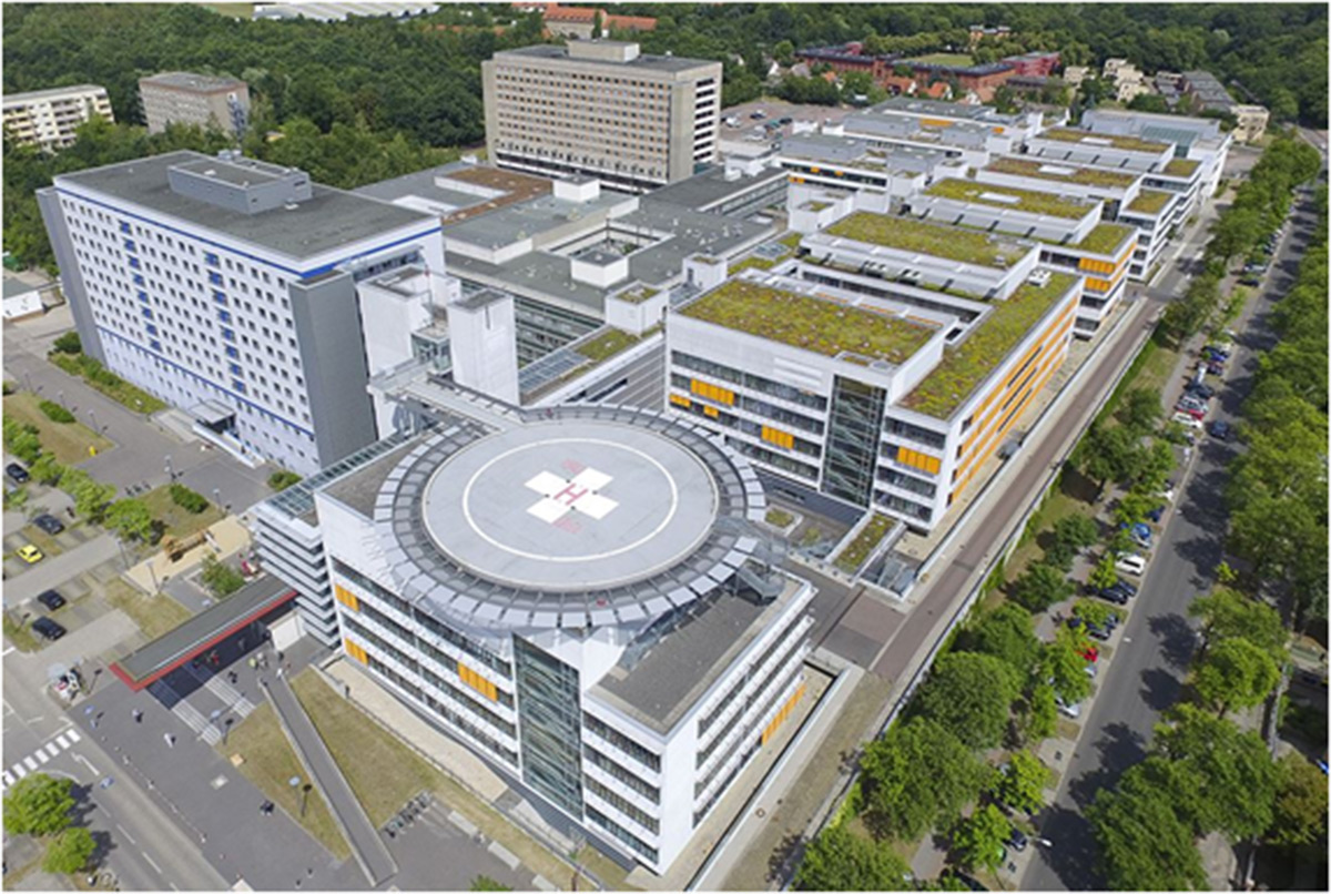Luftaufnahme eines großen Krankenhauskomplexes mit mehreren miteinander verbundenen Gebäuden, einem Hubschrauberlandeplatz mit rotem Kreuz, begrünten Dächern und umliegenden Bäumen und Straßen.