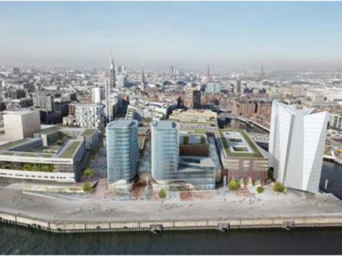 Luftaufnahme einer modernen Stadtlandschaft am Wasser mit drei Glasgebäuden, einer weißen, kantigen Struktur und angrenzenden Straßen mit umliegender Bebauung.