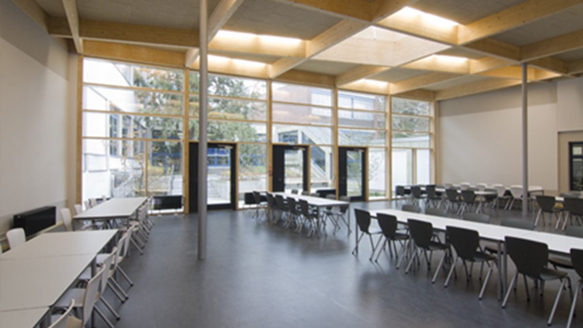 Geräumige Cafeteria mit schwarz-weißen Stühlen, rechteckigen Tischen, großen Fenstern und natürlichem Lichteinfall.