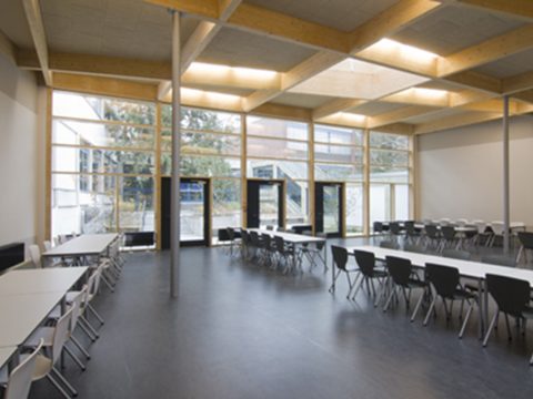 Geräumige Cafeteria mit schwarz-weißen Stühlen, rechteckigen Tischen, großen Fenstern und natürlichem Lichteinfall.