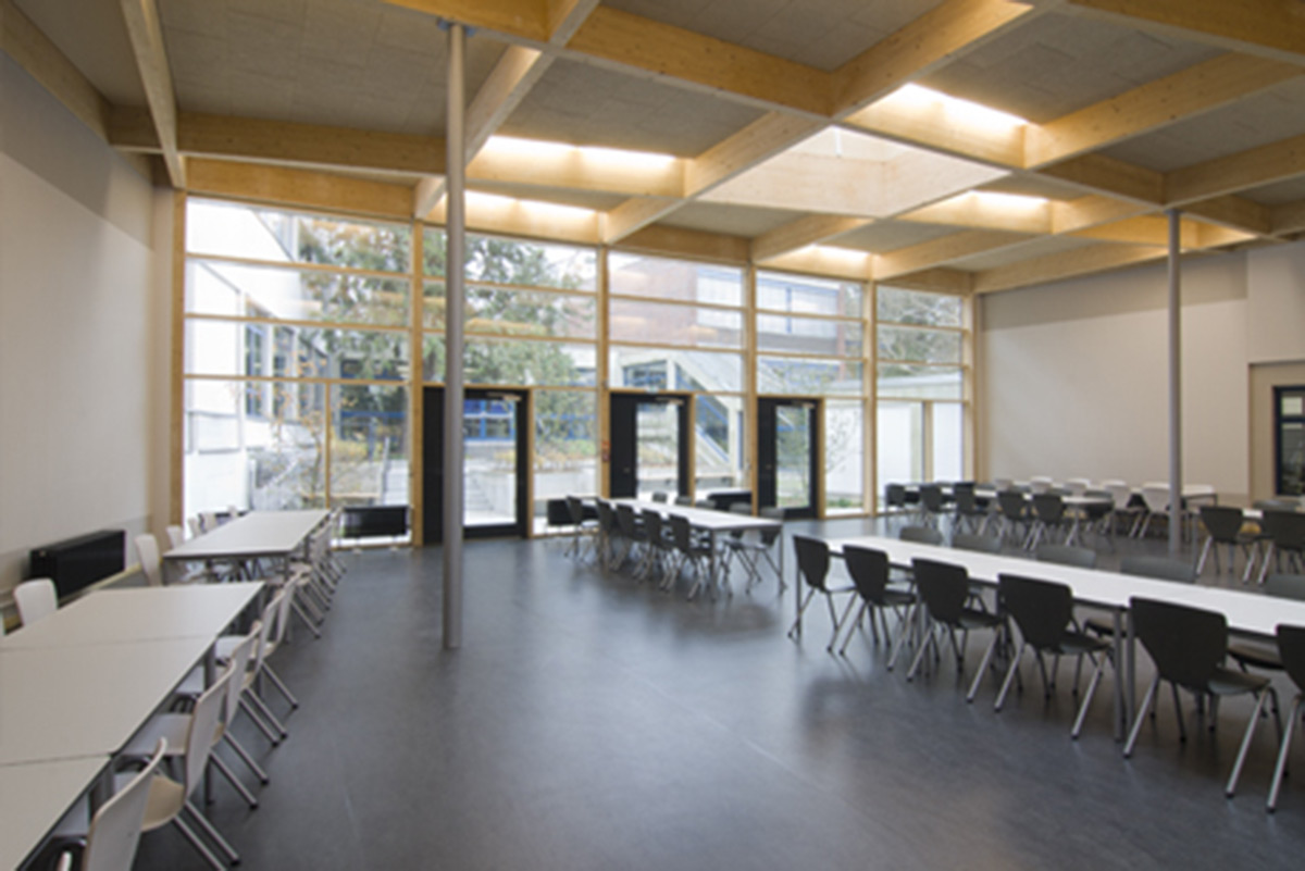 Geräumige Cafeteria mit schwarz-weißen Stühlen, rechteckigen Tischen, großen Fenstern und natürlichem Lichteinfall.