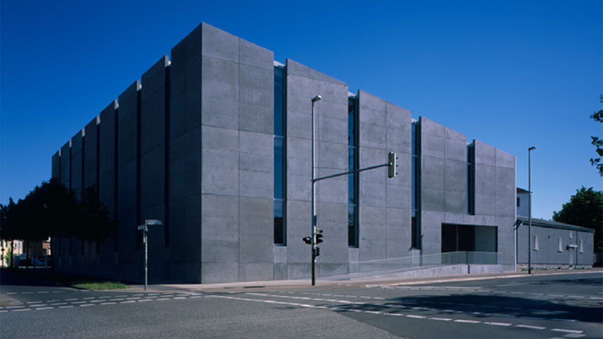 Ein großes, rechteckiges Betongebäude mit schmalen, vertikalen Fenstern steht an einer Straßenecke unter einem strahlend blauen Himmel.