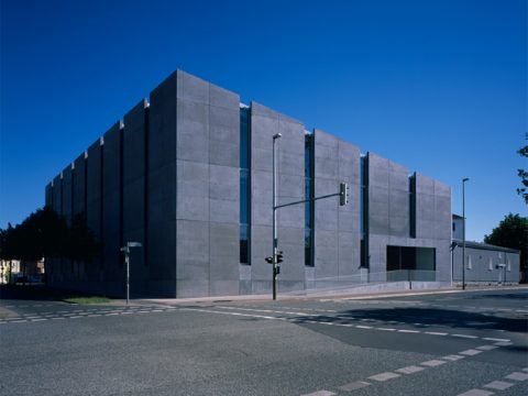 Ein großes, rechteckiges Betongebäude mit schmalen, vertikalen Fenstern steht an einer Straßenecke unter einem strahlend blauen Himmel.