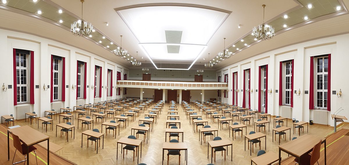 Großer, gut beleuchteter Prüfungssaal mit Reihen von Tischen und Stühlen, Holzfußboden, hoher Decke, Kronleuchtern und hohen Fenstern an den Seiten.