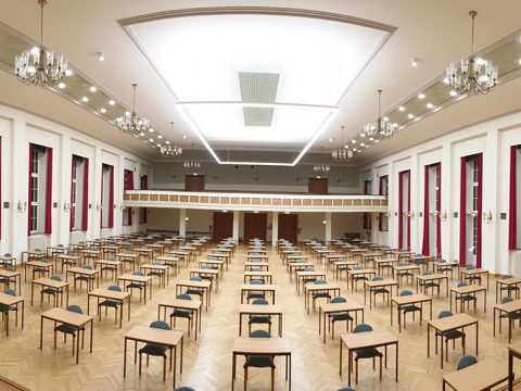 Großer, gut beleuchteter Prüfungssaal mit Reihen von Tischen und Stühlen, Holzfußboden, hoher Decke, Kronleuchtern und hohen Fenstern an den Seiten.
